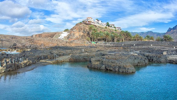 Steile Felsklippen an der Westküste Gran Canarias mit Blick auf den Atlantik. Im Vordergrund ragt eine Aussichtsplattform („Mirador del Balcón“) spektakulär über den Abgrund hinaus, auf der zwei Personen stehen. Darunter brechen die Wellen des Meeres an der felsigen Küste, während sich die zerklüfteten Klippen in die Ferne ziehen.